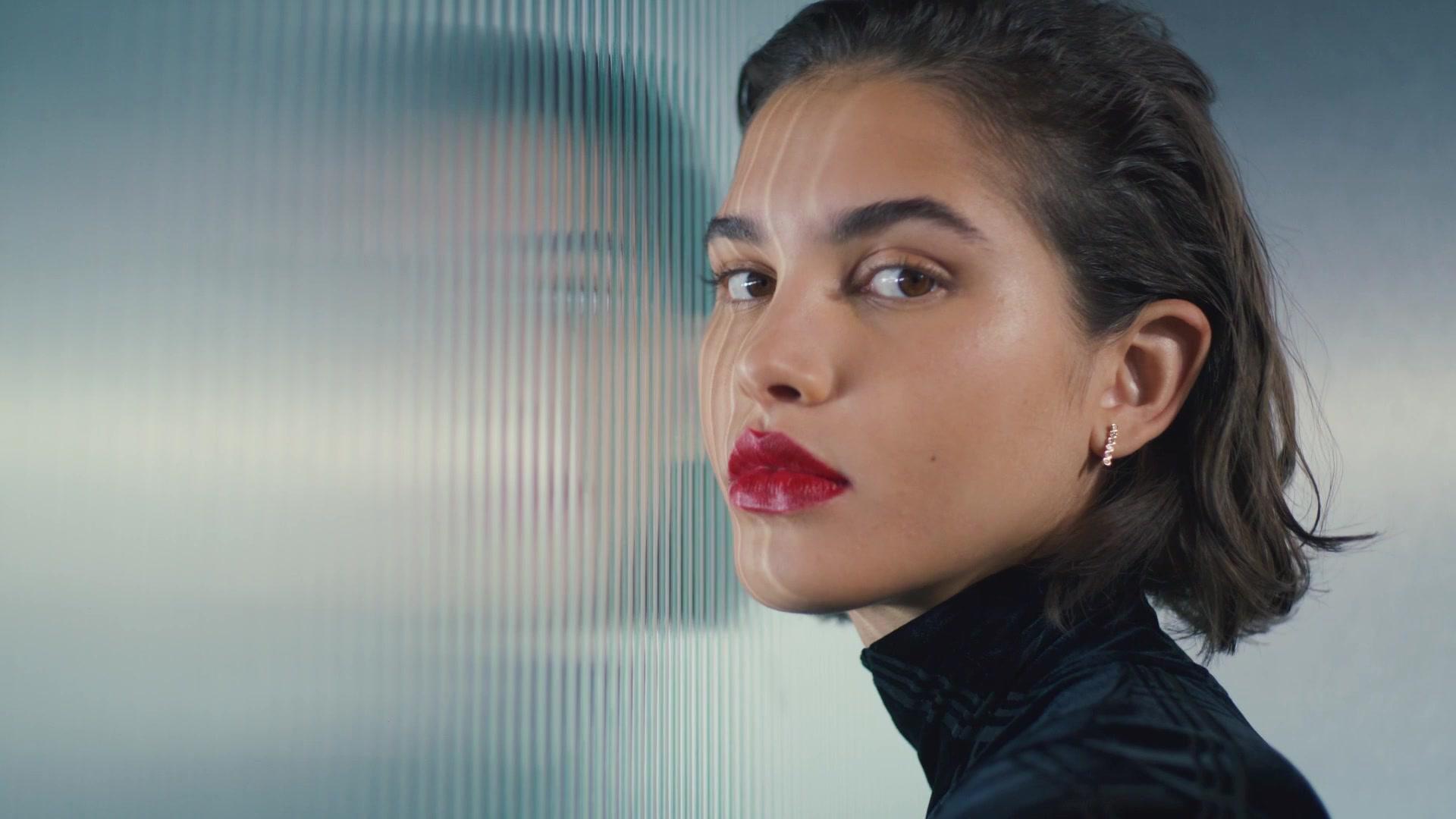 Close portrait of a person with short dark hair and red lipstick against ribbed glass